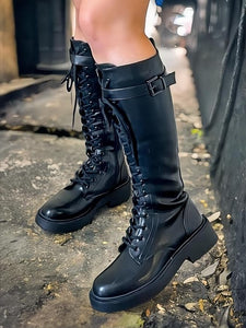 A pair of black knee-high combat boots with lace-up front and decorative buckle detail on a textured ground.