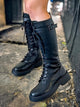 A pair of black knee-high combat boots with lace-up front and decorative buckle detail on a textured ground.